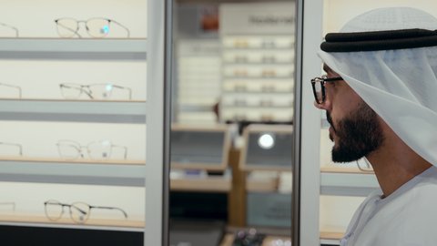زوجان اماراتيان في متجر النظارات، سيده اماراتيه ترتدي عباءه ورديه تستمتع باختيار وقياس نظارة شمسيه مناسبة برفقه زوجها،  رجل عربي خليجي اماراتي يرتدي الكندوره ونظاره طبيه يقوم باختيار نظاره شمسيه، ارفف عرض النظارات ، التسوق وشراء الاكسسورات