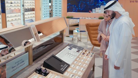 تبادل الاراء بين زوجان اماراتيان في متجر النظارات، سيده اماراتيه ترتدي عباءه ورديه تستمتع باختيارنظاره طبيه مناسبة لزوجها، رجل عربي خليجي اماراتي يرتدي الكندوره يقوم بارتداء نظاره طبيه، ارفف عرض النظارات، التسوق وشراء الاكسسورات