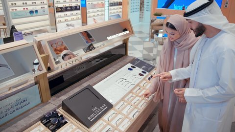 المشاركه بين الازواج في متجر النظارات، سيده اماراتيه ترتدي عباءه ورديه تستمتع باختيارنظاره طبيه مناسبة لزوجها، رجل عربي خليجي اماراتي يرتدي الكندوره يقوم بارتداء نظاره طبيه، ارفف عرض النظارات، التسوق وشراء الاكسسورات