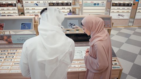 الاختيار بين الازواج في متجر النظارات، زوجان عربيان خليجيان سعوديان يقومان بالتسوق في محل النظارات، ارفف عرض النظارات، التسوق وشراء الاكسسورات في مولات الامارات، زوجان اماراتيان يستمتعان في التسوق، ايماءات السرور الاعجاب