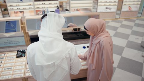 رجل اماراتي يرتدي الكندوره والغتره البيضاء يقوم بقياس النظارة الطبيه المناسبة في محل نظارات برفقه زوجته، الاهتمام بالمظهر الخارجي، التسوق وشراء النظارات الطبيه من المولات، الركن الخاص بالنظارات الطبيه الفخمه، تخفيضات وحسومات