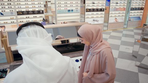 الاختيار بين الازواج في متجر النظارات، زوجان عربيان خليجيان سعوديان يقومان بالتسوق في محل النظارات، ارفف عرض النظارات، التسوق وشراء الاكسسورات في مولات الامارات، زوجان اماراتيان يستمتعان في التسوق، ايماءات السرور الاعجاب
