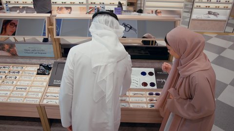 رجل اماراتي يرتدي الكندوره والغتره البيضاء يقوم بقياس النظارة الطبيه المناسبة في محل نظارات برفقه زوجته، الاهتمام بالمظهر الخارجي، التسوق وشراء النظارات الطبيه من المولات، الركن الخاص بالنظارات الطبيه الفخمه، تخفيضات وحسومات