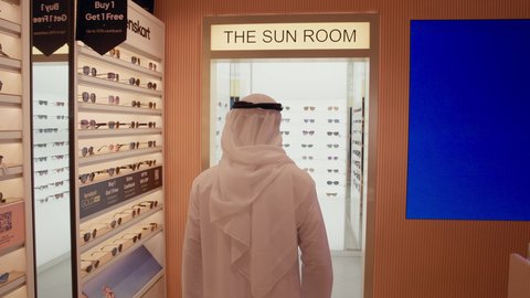 تصوير من الخلف لرجل اماراتي يرتدي الكندوره والغتره البيضاء يقوم بقياس النظارة الشمسيه المناسبة في محل نظارات، الاهتمام بالمظهر الخارجي، التسوق وشراء النظارات الشمسيه من المولات، الركن الخاص بالنظارات الشمسيه الفخمه، تخفيضات وحسومات