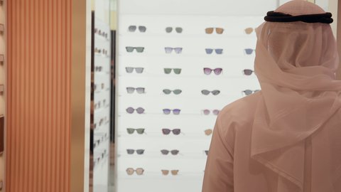 تصوير من الخلف لرجل اماراتي يرتدي الكندوره والغتره البيضاء يقوم بقياس النظارة الشمسيه المناسبة في محل نظارات، الاهتمام بالمظهر الخارجي، التسوق وشراء النظارات الشمسيه من المولات، الركن الخاص بالنظارات الشمسيه الفخمه، تخفيضات وحسومات