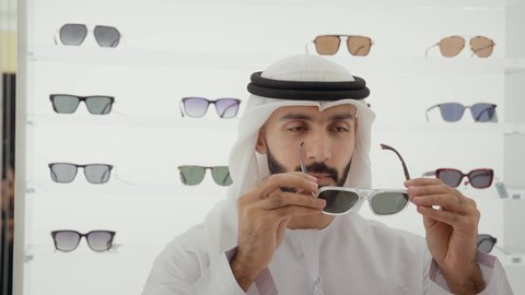 Close-up of an Emirati man wearing a white kandura and ghutra buying suitable sunglasses at an optical store. Taking care of one's appearance, shopping and buying sunglasses from malls.