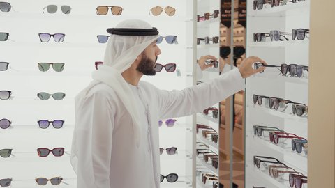 Emirati young man wearing white kandura and ghutra trying on sunglasses in an optician's shop, taking care of his appearance, shopping and buying sunglasses from malls