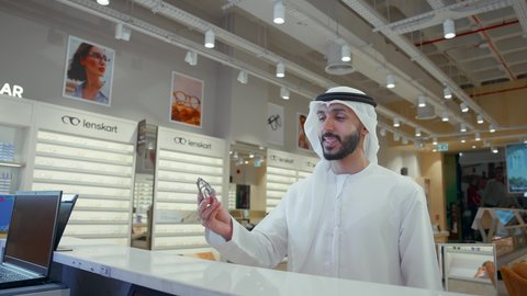 شاب اماراتي يرتدي الكندوره والغتره البيضاء يقوم بشراء النظارة الطبيه المناسبة في محل نظارات ويحملها في كيس ورقي ملون, تحسين الرؤية ومحاولة تعديل البصر، الاهتمام بالمظهر الخارجي، شراء النظارات الخاصة بضعف النظر، التسوق وشراء النظارات الطبيه