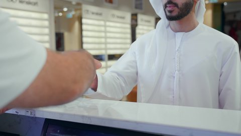 شاب اماراتي يرتدي الكندوره والغتره البيضاء يقوم بشراء النظارة الطبيه المناسبة في محل نظارات ويحملها في كيس ورقي ملون, تحسين الرؤية ومحاولة تعديل البصر، الاهتمام بالمظهر الخارجي، شراء النظارات الخاصة بضعف النظر، التسوق وشراء النظارات الطبيه