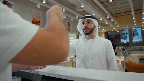 رجل اماراتي يرتدي الكندوره والغتره البيضاء يقوم بشراء النظارة الشمسيه المناسبة في محل نظارات، الاهتمام بالمظهر الخارجي، التسوق وشراء النظارات الشمسيه من المولات، شاب اماراتي يقوم بحمل كيس ورقي ملون