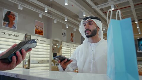 إتمام عملية الدفع باستخدام الهاتف المحمول، تطبيقات التسوق والشراء الالكتروني عبر الهاتف، اقتناء وشراء هدايا من احدي المولات الاماراتيه، رجل عربي خليجي اماراتي يحمل كيس ورقي ملون