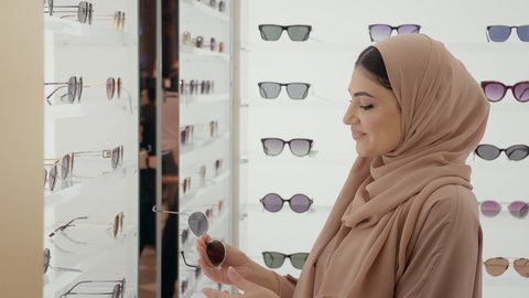 التسوق وشراء النظارات الشمسيه من المولات، الركن الخاص بالنظارات الشمسيه الفخمه، تصوير مقرب لامراة عربيه خليجيه اماراتية محجبه ترتدي العباءه الملونة تقوم بقياس النظارة الشمسيه المناسبة في محل نظارات، الاهتمام بالمظهر الخارجي، تخفيضات وحسومات