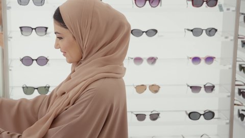 الركن الخاص بالنظارات الشمسيه الفخمه، التسوق وشراء النظارات الشمسيه من المولات، تصوير مقرب لامراة عربيه خليجيه اماراتية محجبه ترتدي العباءه الملونة تقوم بقياس النظارة الشمسيه المناسبة في محل نظارات، الاهتمام بالمظهر الخارجي، تخفيضات وحسومات