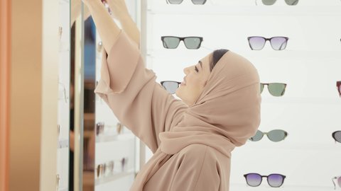 التسوق وشراء النظارات الشمسيه من المولات، الركن الخاص بالنظارات الشمسيه الفخمه، تصوير مقرب لامراة عربيه خليجيه اماراتية محجبه ترتدي العباءه الملونة تقوم بقياس النظارة الشمسيه المناسبة في محل نظارات، الاهتمام بالمظهر الخارجي، تخفيضات وحسومات