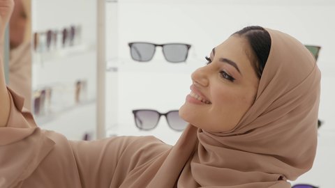 الركن الخاص بالنظارات الشمسيه الفخمه، تصوير مقرب لامراة عربيه خليجيه اماراتية محجبه ترتدي العباءه الملون تقوم بقياس النظارة الشمسيه المناسبة في محل نظارات، لاهتمام بالمظهر الخارجي، التسوق وشراء النظارات الشمسيه من المولات، تخفيضات وحسومات