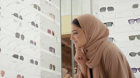 الاهتمام بالمظهر الخارجي، التسوق وشراء النظارات الشمسيه من المولات،تصوير مقرب لامراة عربيه خليجيه اماراتية محجبه ترتدي العباءه الملون تقوم بقياس النظارة الشمسيه المناسبة في محل نظارات