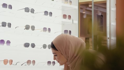 التسوق وشراء النظارات الشمسيه من المولات،تصوير مقرب لامراة عربيه خليجيه اماراتية محجبه ترتدي العباءه الملون تقوم بقياس النظارة الشمسيه المناسبة في محل نظارات، الاهتمام بالمظهر الخارجي