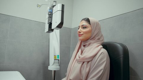 مفهوم الخدمات والرعاية الصحية في المستشفيات والمراكز، تقييم الرؤية والكشف عن أمراض العيون للمريض، استخدام الأجهزة الحديثة المتطورة،تصوير مقرب  من الجانب لامراة عربيه خليجيه اماراتية ترتدي العباءه الملونه في عيادة مختصة لفحص العيون تنظر من خلال مجموعة من العدسات