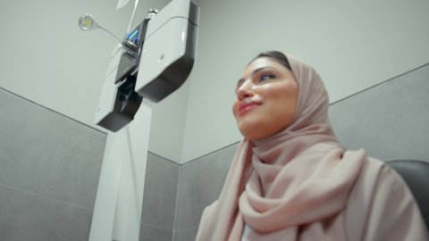 مفهوم الخدمات والرعاية الصحية في المستشفيات والمراكز، تقييم الرؤية والكشف عن أمراض العيون للمريض، استخدام الأجهزة الحديثة المتطورة،تصوير مقرب  من الجانب لامراة عربيه خليجيه اماراتية ترتدي العباءه الملونه في عيادة مختصة لفحص العيون تنظر من خلال مجموعة من العدسات