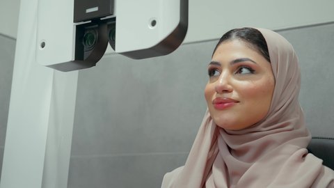 تقييم الرؤية والكشف عن أمراض العيون للمريض، استخدام الأجهزة الحديثة المتطورة،تصوير مقرب لامراة عربيه خليجيه اماراتية ترتدي العباءه الملونه في عيادة مختصة لفحص العيون تنظر من خلال مجموعة من العدسات ،مفهوم الخدمات والرعاية الصحية في المستشفيات والمراكز