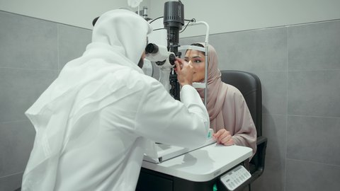 تقييم مدى الرؤية والكشف عن الامراض العينية، مفهوم الخدمات والرعاية الصحية في المشافي والمراكز الطبية، تشخيص وتحديد المشاكل البصرية، استخدام الأجهزة الحديثة المتطورة، تصوير مقرب من الخلف لطبيب عربي خليجي اماراتي يقوم بفحص عينى امراه  اماراتية بجهاز المنظار العيني