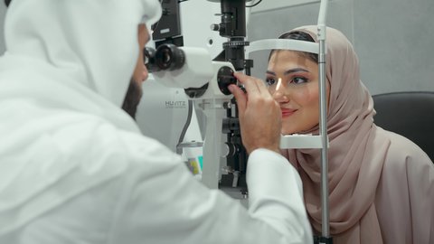 The concept of healthcare services in hospitals and medical centers, diagnosis and identification of visual problems, use of modern advanced equipment, close-up of an Emirati Gulf Arab doctor examining the eyes of an Emirati woman with an ophthalmoscope, assessing vision and detecting eye diseases.