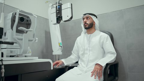 Periodic visits to eye clinics in the Emirates, the concept of services and health care in hospitals and centers, the use of modern advanced equipment, an Arab Gulf Emirati man wearing traditional clothing in a specialized eye examination clinic looking through a set of lenses