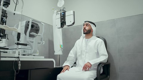 مفهوم الخدمات والرعاية الصحية في المستشفيات والمراكز، استخدام الأجهزة الحديثة المتطورة،رجل عربي خليجي اماراتي يرتدي الزي التقليدي في عيادة مختصة لفحص العيون ينظر من خلال مجموعة من العدسات ، الزيارة الدورية للعيادات العينية في الامارات