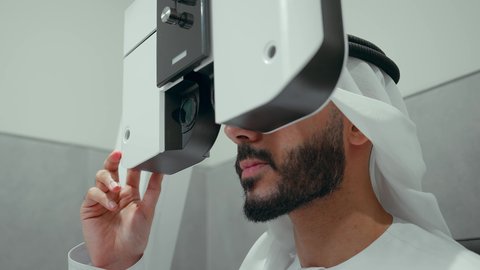 استخدام الأجهزة الحديثة المتطورة، تصوير مقرب من الجانب لرجل عربي خليجي اماراتي يرتدي الزي التقليدي في عيادة مختصة لفحص العيون ينظر من خلال مجموعة من العدسات ،مفهوم الخدمات والرعاية الصحية في المستشفيات والمراكز، الزيارة الدورية للعيادات العينية في الامارات