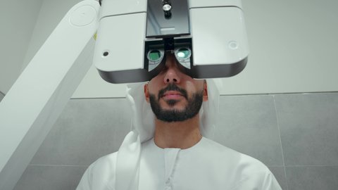 استخدام الأجهزة الحديثة المتطورة، تقييم الرؤية والكشف عن أمراض العيون للمريض،تصوير مقرب لرجل عربي خليجي اماراتي يرتدي الزي التقليدي في عيادة مختصة لفحص العيون ينظر من خلال مجموعة من العدسات ،مفهوم الخدمات والرعاية الصحية في المستشفيات والمراكز، الزيارة الدورية للعيادات العينية في الامارات