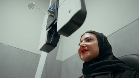 Diagnosing and identifying visual problems, using modern advanced equipment, a close-up side view of an Emirati Gulf Arab woman wearing an abaya in front of an eye examination device to ensure healthy vision, assessing vision and detecting eye diseases, the concept of services and healthcare in hospitals and centers.