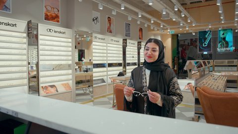 الاهتمام بالمظهر الخارجي، امراه عربيه خليجيه اماراتية محجبه ترتدي العباءه السوداء تقوم بشراء النظارة الشمسيه المناسبة في محل نظارات بايماءات السعاده و السرور، شراء النظارات الخاصة بحماية النظر، التسوق وشراء النظارات الشمسية