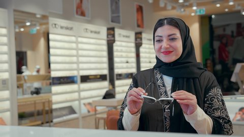 التسوق وشراء النظارات الشمسية، شراء النظارات الخاصة بحماية العين من الاشعه، الاهتمام بالمظهر الخارجي، تصوير مقرب لامراه عربيه خليجيه اماراتية محجبه ترتدي العباءه السوداء تقوم بشراء النظارة الشمسيه المناسبة في محل نظارات بايماءات السعاده و السرور