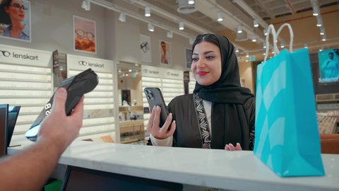 إتمام عملية الدفع باستخدام الهاتف المحمول، تطبيقات التسوق والشراء الالكتروني عبر الهاتف، اقتناء وشراء هدايا من احدي المولات الاماراتيه، امراة عربيه خليجيه اماراتية ترتدي العباءه السوداء تحمل كيس ورقي ملون بايماءات السعاده و السرور
