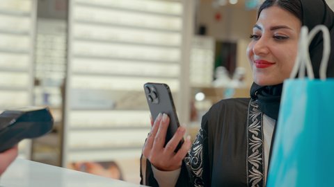 تطبيقات التسوق والشراء الالكتروني عبر الهاتف، إتمام عملية الدفع باستخدام الهاتف المحمول، اقتناء وشراء هدايا من احدي المولات الاماراتيه، امراة عربيه خليجيه اماراتية ترتدي العباءه السوداء تحمل كيس ورقي ملون بايماءات السعاده و السرور