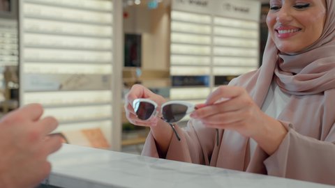Caring about one's appearance, a veiled Emirati Gulf Arab woman buying suitable sunglasses in an optical store carrying them in a colorful paper bag with gestures of happiness and pleasure, improving vision and trying to correct eyesight, buying glasses for weak eyesight, shopping and buying prescription glasses