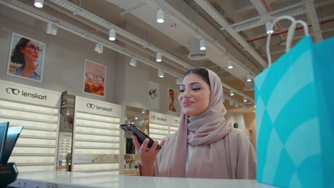 Mobile shopping and purchasing applications, completing the payment process using a mobile phone, purchasing and buying gifts from one of the Emirati malls, an Emirati Gulf Arab woman wearing a colorful abaya carrying a colorful paper bag with gestures of happiness and pleasure