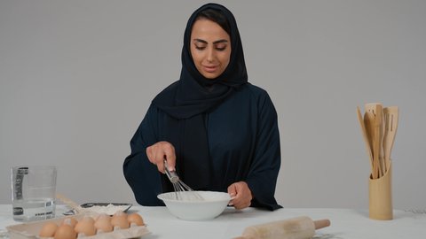 تعابير الموافقه و القبول، إعداد المخبوزات والمعجنات الشهية، أدوات خاصة بالمطبخ، مسؤولية ربة المنزل، تجهيز المكونات لصناعة الخبز، تصوير مقرب لامرأة عربية خليجية اماراتية محجبة ترتدي العباءه السوداء تحمل بيدها الخفاقة اليدوية تقوم بتحضير العجين بايماءات السعاده و السرور