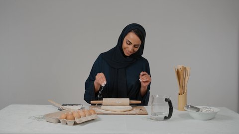 Kitchen tools, using a dough rolling tool, close-up of a veiled Emirati Gulf Arab woman wearing a black abaya sprinkling flour on the dough, preparing ingredients for making bread, preparing delicious baked goods and pastries, the responsibility of the housewife
