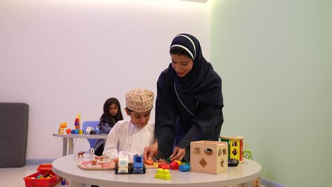 معلمة لرياض أطفال عربية عمانيه تختص بالاعتناء بالأطفال، أداء الكثير من الأنشطة الممتعة، محاوله ادخال القطع في مكانها الصحيح ، طفل عربي خليجي عماني يرتدي الدشداشه والكمه في رياض الأطفال والحاضنات