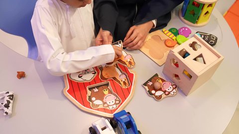 معلمه عربيه خليجيه عمانيه تقوم بمساعده الطفل لوضع البزل في مكانها الصحيح، طفل عربي خليجي عماني يرتدي الدشداشه والكمه يقوم بتركيب الاحجيه، ايماءات يدين تدل علي الحفاوه والتشجيع، مهارات تعليميه في رياض الاطفال، مجموعه من العاب الذكاء علي الطاوله