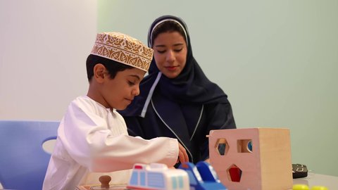 أداء الكثير من الأنشطة الممتعة،  معلمة لرياض أطفال عربية عمانيه تختص بالاعتناء بالأطفال، طفل عربي خليجي عماني يرتدي الدشداشه والكمه في رياض الأطفال والحاضنات، محاوله ادخال القطع في مكانها الصحيح