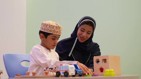 An Omani Arab kindergarten teacher specializing in taking care of children, performing many fun activities, an Omani child wearing a dishdasha and a kuma in kindergartens and nurseries, an Omani Arab Gulf child trying to put pieces in their correct place