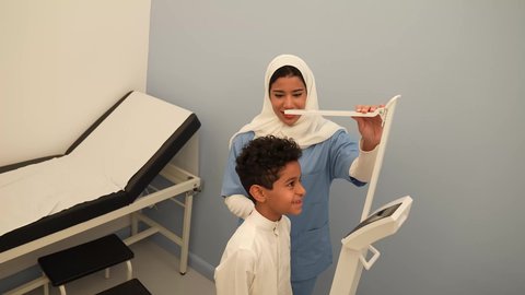 ممرضه عربية خليجية سعوديه تقوم بقياس الطول للطفل، ولد سعودي يرتدي الزي السعودي في موعد مع الطبيبه لعمل الفحوصات اللازمه، مفهوم الطب والرعاية الصحية بالأطفال، الفحص الطبي للأطفال، الرعاية الطبية للأطفال، ادوات طبيه متقدمه للقياس