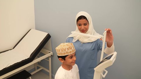 ولد عماني يرتدي الدشداشه والكمه في موعد مع الطبيبه لعمل الفحوصات اللازمه، مفهوم الطب والرعاية الصحية بالأطفال، الفحص الطبي  والرعاية الطبية ،  طبيبه عربية خليجية عمانيه تقوم بقياس الطول للطفل،  ادوات طبيه متقدمه للقياس