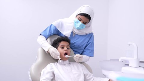 استخدام الأدوات الطبية السنية المعقمة،تصوير مقرب لفتي عربي خليجي سعودي في عيادة سنية،  طبيبه أسنان عربيه خليجيه سعوديه ترتدي اليونيفورم والقفازات والكمامة الطبية تمسك بيدها ادوات طبية تقوم بتنظيف اسنان الطفل، مفهوم العناية بأسنان الأطفال