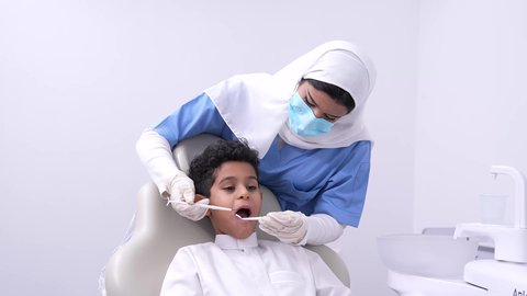 مفهوم العناية بأسنان الأطفال ، فتي عربي خليجي سعودي في عيادة سنية يتلقى العلاج اللازم، استخدام الأدوات الطبية السنية المعقمة، طبيبه أسنان عربيه خليجيه سعوديه ترتدي اليونيفورم والقفازات والكمامة الطبية تمسك بيدها ادوات طبية تقوم بتنظيف اسنان الطفل