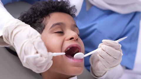 تصوير مقرب لفتي عربي خليجي سعودي في عيادة سنية، استخدام الأدوات الطبية السنية المعقمة، طبيبه أسنان عربيه خليجيه سعوديه ترتدي اليونيفورم والقفازات والكمامة الطبية تمسك بيدها ادوات طبية تقوم بتنظيف اسنان الطفل، مفهوم العناية بأسنان الأطفال