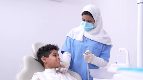 طبيبه أسنان عربيه خليجيه سعوديه ترتدي اليونيفورم والقفازات والكمامة الطبية تمسك بيدها ادوات طبية تقوم بتنظيف اسنان الطفل، فتي عربي خليجي سعودي في عيادة سنية يتلقى العلاج اللازم، استخدام الأدوات الطبية السنية المعقمة، مفهوم العناية بأسنان الأطفال