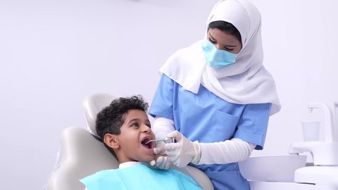 A Saudi Arabian Gulf boy in a dental clinic receiving the necessary treatment, using sterile dental medical tools, a Saudi Arabian Gulf female dentist wearing a uniform, gloves and a medical mask holding medical tools in her hand, the concept of children's dental care